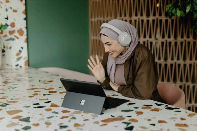 A smiling woman wearing a hijab and headphones waving at her laptop during a video call.